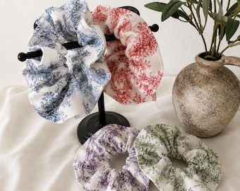 May include: A collection of fabric scrunchies in various colors, including blue, red, purple, and green, each adorned with a delicate floral pattern. Some scrunchies are displayed on a black stand, while others rest on a white surface.