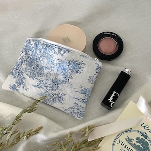 Może przedstawiać: Biało-niebieska kosmetyczka z zamkiem błyskawicznym, z małym kompaktem, okrągłym pudrem do policzków i czarną szminką. Kosmetyczka leży na białym lnianym materiale z suszonymi źdźbłami owsa. W prawym dolnym rogu zdjęcia znajduje się mała karta z tekstem "yusunnyday HANDMADE".