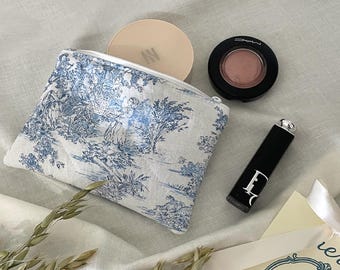 May include: A white and blue toiletry bag with a zipper, containing a compact, blush, and lipstick. The bag features a scenic design. A tag with the text "yusunnyday HANDMADE" is also present.
