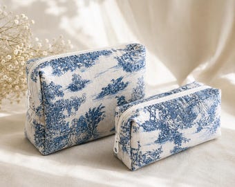 May include: Two toiletry bags in blue and white. The bags feature a classic toile pattern with trees and landscapes. The bags have white zippers and are made of a textured fabric. One bag is larger than the other.
