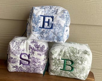May include: Three toile-patterned zippered pouches with embroidered initials. The pouch on the top is blue and white with the letter "E" embroidered in blue. The pouch in the middle is purple and white with the letter "S" embroidered in purple. The pouch on the bottom is green and white with the letter "B" embroidered in green.