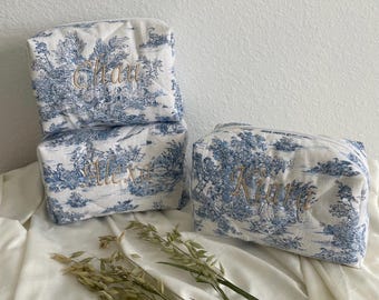 May include: Three white toiletry bags with a blue landscape pattern. Each bag has a different name embroidered in gold thread: "Chan", "Alexa", and "Kiara". The bags are rectangular and quilted.