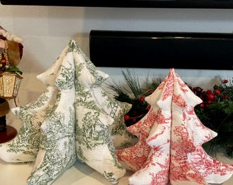 May include: Three fabric Christmas tree decorations. Two trees are white with a green toile pattern, and one is red with a white toile pattern. The trees are arranged on a white surface, with greenery in the background.