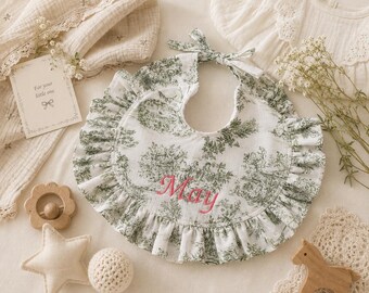 May include: A white baby bib with a green toile pattern and a ruffled edge. The bib has the name "May" embroidered in pink. A card with the text "For your little one" is also visible.