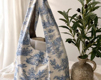 May include: A blue and white toile print tote bag with a wide opening and a single handle. The bag is made of fabric and has a classic, pastoral design. A vase with olive branches is in the background.