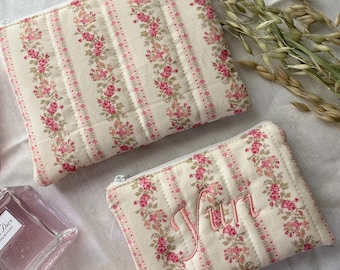 May include: Two floral print cosmetic bags with a cream background and pink roses. One bag has the name "Yun" embroidered in pink. The bags have a quilted design and a zipper closure. The bags are rectangular in shape.