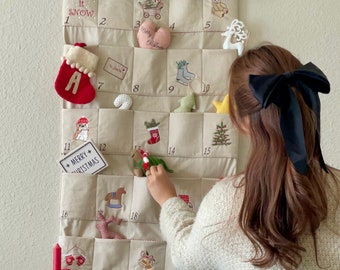 May include: A beige fabric advent calendar with embroidered numbers and Christmas-themed illustrations. The calendar has pockets for small gifts or treats. A person is reaching into one of the pockets.