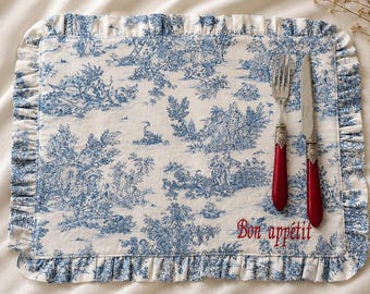 May include: A white and blue toile placemat with a ruffled edge, featuring a pastoral scene. Silverware with red handles rests on the placemat. The embroidered text "Bon appétit" is visible.