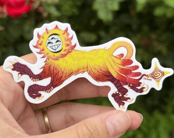 May include: A yellow and red sticker of a mythical creature with a sun-like head and three eyes. The creature has a long tail with a spiral design and the text "CYTKAKI" is visible on the tail.
