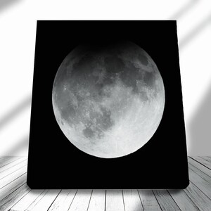 May include: A black square canvas displays a detailed image of a lunar eclipse. The moon is mostly gray with dark shadows, showing craters and textures. The canvas is set against a white wall and a wooden floor.