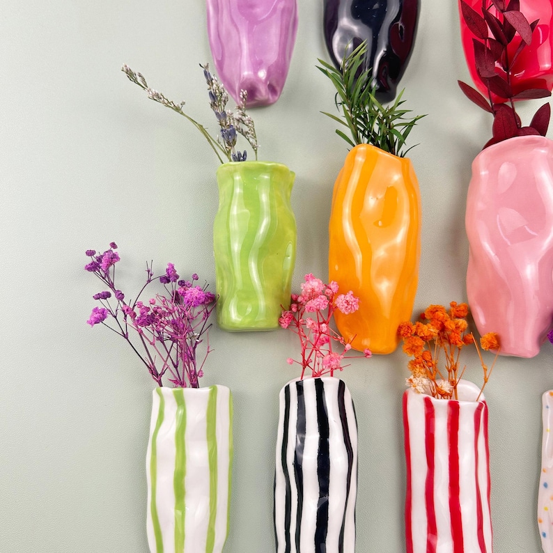 May include: A collection of colorful ceramic vases in various shapes and sizes, some with vertical stripes. Each vase holds dried flowers and greenery. The vases are in shades of green, orange, pink, and red.