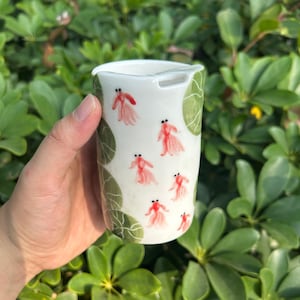 May include: A white ceramic travel mug with a lid, featuring a hand-painted design of red goldfish and green leaves. The mug is held against a backdrop of green foliage, suggesting a nature-inspired theme.