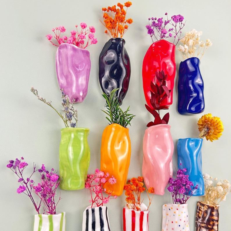 May include: A collection of colorful ceramic vases in various shapes and sizes, each holding dried flowers and greenery. The vases are in shades of pink, green, orange, red, blue, and black, with some featuring striped and dotted patterns. A decorative floral arrangement.