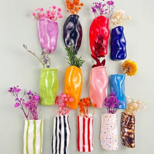 May include: A collection of colorful ceramic wall vases in various shapes and sizes, each holding dried flowers. Vases are in shades of pink, green, orange, blue, and white, with striped and dotted designs.