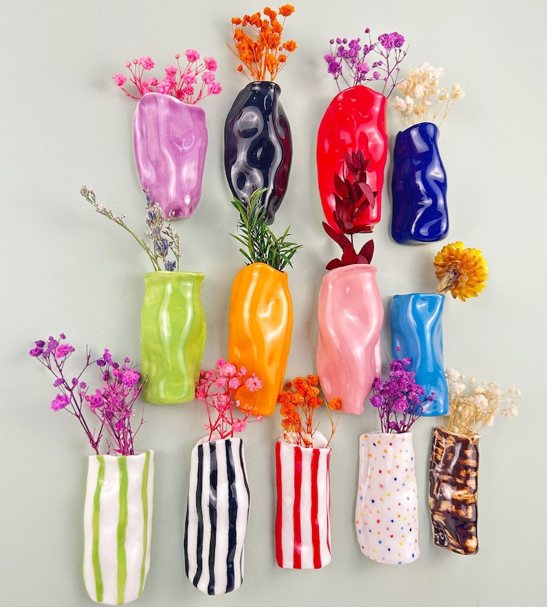 May include: A collection of colorful ceramic wall vases in various shapes and sizes, each holding dried flowers. Vases are in shades of pink, green, orange, blue, and white, with striped and dotted designs.