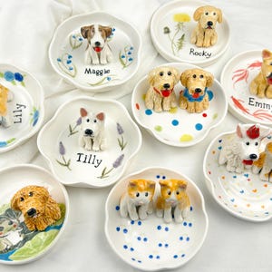 May include: A collection of ceramic dog-themed trinket dishes. Each dish features a different breed of dog in a 3D design, with names like "Maggie", "Rocky", and "Lily" written on the dish. The dishes are white with colorful accents.