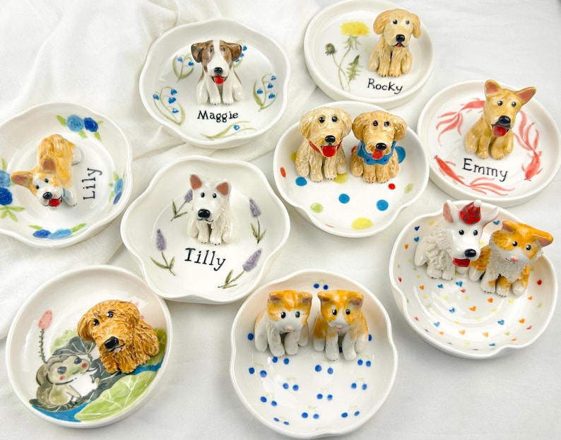 May include: A collection of ceramic dog-themed trinket dishes. Each dish features a different breed of dog in a 3D design, with names like "Maggie", "Rocky", and "Lily" written on the dish. The dishes are white with colorful accents.