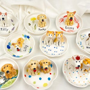 May include: A collection of small, white ceramic dishes, each adorned with a hand-painted dog figurine. The dishes feature various dog breeds and are decorated with floral and dot patterns. Each dish has a dog's name written on it.