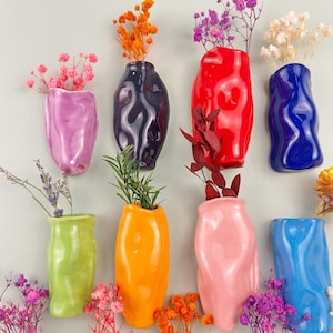 May include: A collection of eight colorful, ceramic wall vases in various shades including pink, purple, black, red, blue, green, and orange. Each vase holds dried flowers or greenery, creating a vibrant display. The vases have a unique, organic shape.