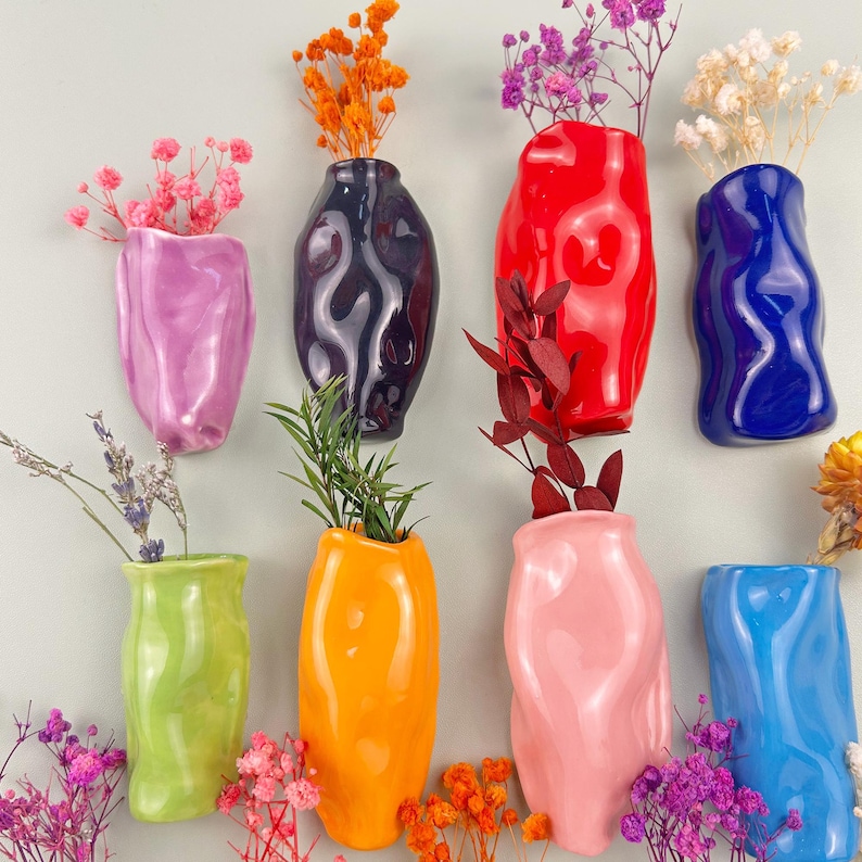 May include: A collection of eight colorful, ceramic wall vases in various shades including pink, purple, black, red, blue, green, and orange. Each vase holds dried flowers or greenery, creating a vibrant display. The vases have a unique, organic shape.