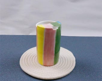 May include: A colorful ceramic mug with vertical stripes of yellow, pink, and green, resembling a large pencil. The mug sits on a round, off-white woven coaster. The background is a solid blue surface and a white backdrop.
