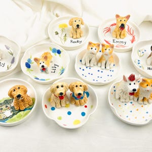 May include: A collection of ceramic dog-themed trinket dishes. Each dish features a different dog breed figurine, with names like "Tilly," "Lily," and "Rocky" written on them. The dishes are white with colorful floral or dot designs.