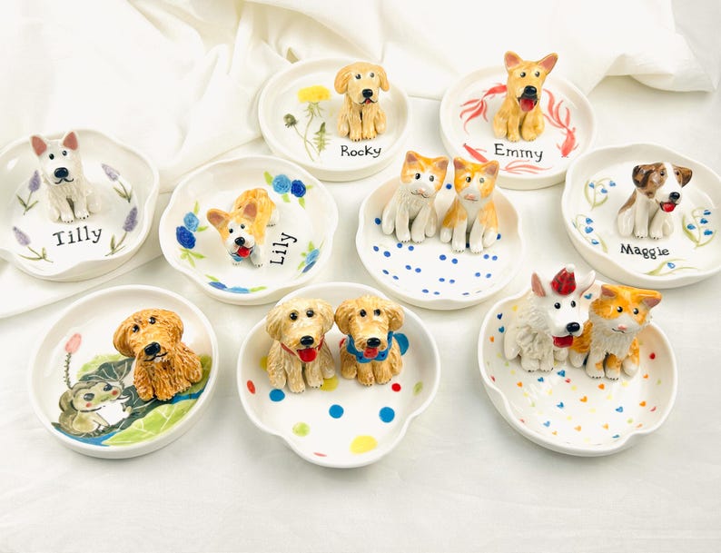 May include: A collection of ceramic dog-themed trinket dishes. Each dish features a different dog breed figurine, with names like "Tilly," "Lily," and "Rocky" written on them. The dishes are white with colorful floral or dot designs.