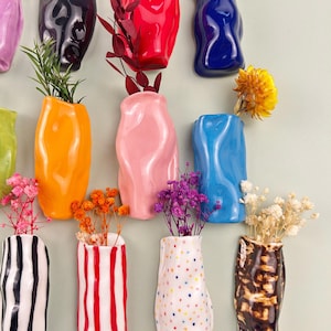 May include: A collection of colorful ceramic vases in various shapes and sizes, each holding dried flowers and greenery. The vases are in shades of orange, pink, blue, and black, with some featuring striped and dotted patterns. The arrangement is against a light green background.