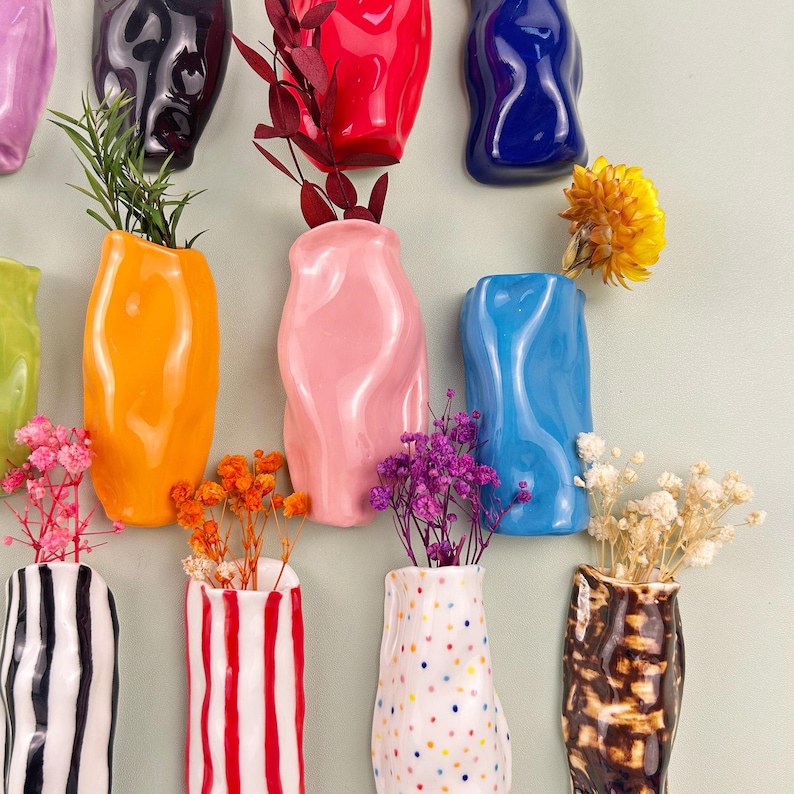 May include: A collection of colorful ceramic vases in various shapes and sizes, each holding dried flowers and greenery. The vases are in shades of orange, pink, blue, and black, with some featuring striped and dotted patterns. The arrangement is against a light green background.