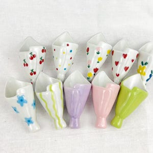 Puede incluir: Una colección de diez reposa palillos de cerámica con forma de pequeños ramos. Las piezas presentan varios diseños, incluyendo cerezas rojas, lunares coloridos, flores rosas y amarillas, corazones rojos, flores azules, rayas verdes y colores pastel sólidos.