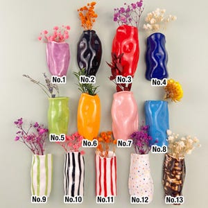 May include: A collection of thirteen ceramic vases in various colors and designs, each holding dried flowers. The vases include solid colors like pink, blue, and orange, as well as striped and dotted patterns. Each vase is labeled with a number.