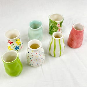 Könnte beinhalten: Eine Sammlung von sieben kleinen, handgefertigten Keramikvasen. Jede Vase hat eine einzigartige Form und ein einzigartiges Design, darunter Blumenmuster, Tupfen und Streifen. Die Vasen sind in verschiedenen Farben wie Grün, Rosa und Weiß auf einer weißen Oberfläche angeordnet.