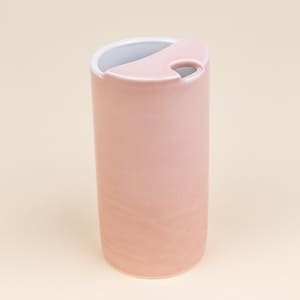 May include: A cylindrical, pink ceramic tumbler with a white interior and a curved, pink lid featuring a small opening. The tumbler has a smooth, matte finish and a minimalist design, suitable for beverages.