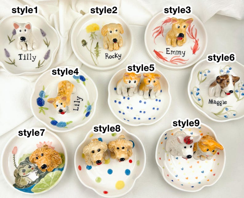 May include: A collection of nine small, white ceramic dishes, each adorned with a 3D dog figurine. The dishes feature various dog breeds and are decorated with floral and dot patterns. Each dish has a dog's name written on it.