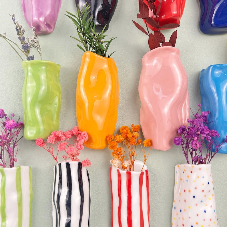 May include: A collection of colorful ceramic vases in various shapes and sizes, some with vertical stripes or polka dots. The vases are filled with dried flowers and greenery, creating a vibrant display. The color palette includes green, orange, pink, and blue.