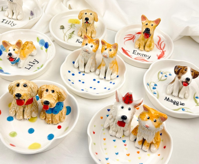 May include: A collection of ceramic dog figurines on small, white, scalloped dishes. Each dish features a different dog breed, with names like "Lily" and "Maggie" written on them. The dishes are decorated with colorful patterns and floral accents.