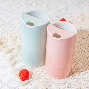 May include: Two ceramic vases, one light blue and one pink, with a unique design. Each vase has a curved top with a small opening. The vases are cylindrical and sit on a white lace surface, with a red berry nearby.