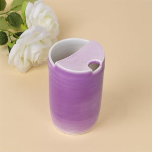 May include: A lavender ceramic mug with a unique curved lid and a small hole. The mug has a white interior and a textured exterior. The design is simple and elegant, suitable for holding beverages.