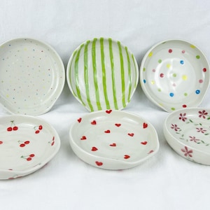 May include: A collection of six white ceramic dishes with various hand-painted designs. The designs include colorful polka dots, green stripes, red cherries, red hearts, and pink floral patterns. Each dish has a unique shape and size.