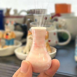 May include: A miniature ceramic vase with a glossy, pale pink and white marbled finish. The small, bulbous vessel features a narrow neck and a flared rim, containing several thin, white, straight bristles extending upward from the opening.