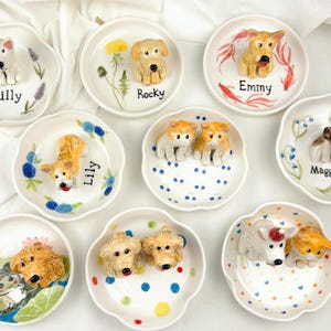 May include: A collection of small, white ceramic dishes, each adorned with a 3D dog figurine. The dishes feature various dog breeds and are decorated with floral or dot patterns. Each dish has a dog's name written on it, such as "Tilly" and "Rocky".