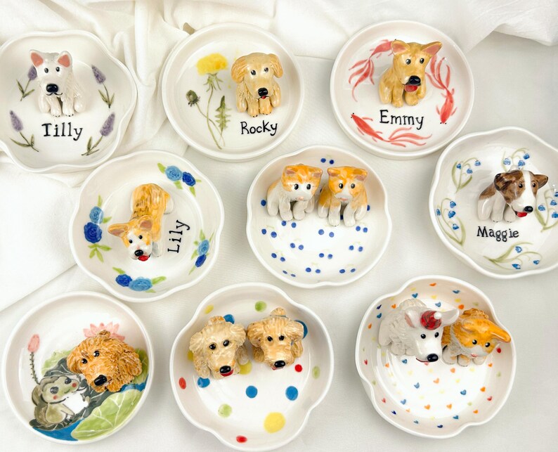 May include: A collection of small, white ceramic dishes, each adorned with a 3D dog figurine. The dishes feature various dog breeds and are decorated with floral or dot patterns. Each dish has a dog's name written on it, such as "Tilly" and "Rocky".