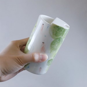 Puede incluir: Taza de viaje de cerámica blanca con tapa, con un diseño caprichoso de nenúfares verdes y pequeñas criaturas rosadas con forma de camarón. La taza tiene un acabado brillante y una estética limpia y moderna.