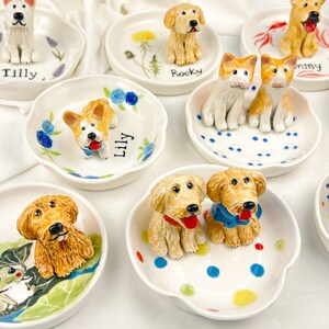 May include: A collection of ceramic dog figurines on small, white, scalloped dishes. Each dish features a different dog breed, with hand-painted details and names like "Tilly" and "Rocky". Some dishes have floral or dot designs.