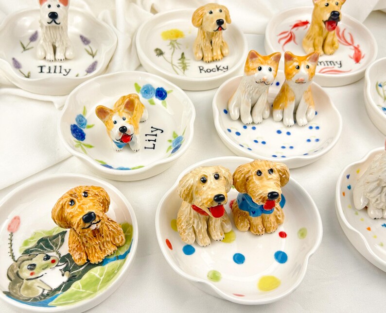 May include: A collection of ceramic dog figurines on small, white, scalloped dishes. Each dish features a different dog breed, with hand-painted details and names like "Tilly" and "Rocky". Some dishes have floral or dot designs.