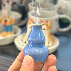 May include: A small, blue, gourd-shaped ceramic vase with a smooth, glossy finish. The vase features a narrow neck that widens into a rounded body, designed to hold a single stem or small decorative element. The surface reflects light evenly.