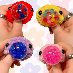 May include: Four plush toys with different colored fur, each with a clear plastic bubble filled with colorful squishy balls. The toys are red, yellow, purple, and pink.