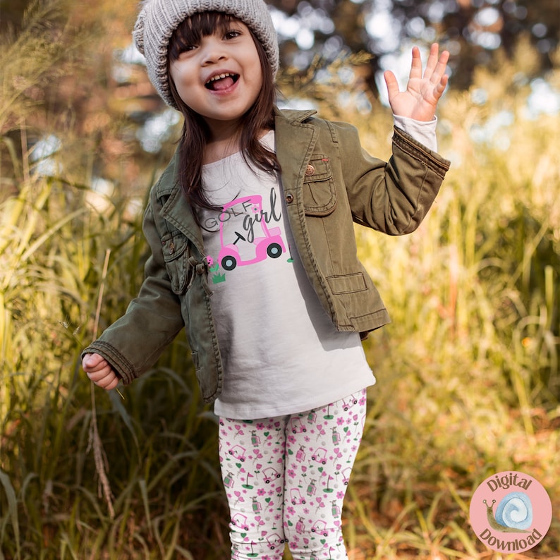 May include: A young girl wearing a white shirt with the text "Golf Girl" and a pink golf cart graphic. She is also wearing a green jacket and pink and white leggings with a golf cart pattern.