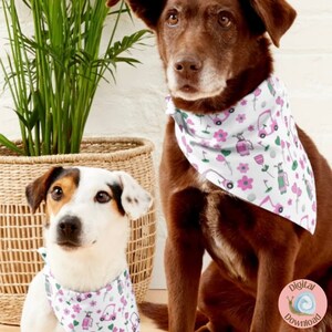 May include: Two dogs wearing white bandanas with a pink and green golf cart pattern. The bandanas have pink flowers, green golf clubs, and white golf balls. The dogs are sitting in front of a wicker basket filled with green plants.