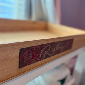 Può includere: Un vassoio di legno con una placca decorativa rossa e dorata che dice "Ruby" sopra.